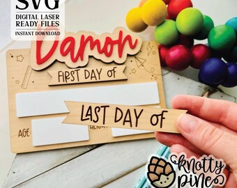 May include: A wooden sign with the name "Damon" in red letters, and the text "First Day of" and "Last Day of" on banners. The sign has spaces for writing in the age and height. The sign is on a wooden surface with pencils and a rainbow colored ball chain.