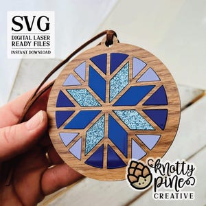 Può includere: Un ornamento in legno con un design a fiocco di neve blu e viola. L'ornamento è rotondo e ha una finitura in legno marrone. Il fiocco di neve è composto da diversi triangoli e ha una texture scintillante. L'ornamento ha una corda per appenderlo.