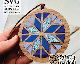 May include: A wooden ornament with a blue and purple snowflake design. The ornament is round and has a brown wood grain finish. The snowflake is made up of multiple triangles and has a glittery texture. The ornament has a string attached to it for hanging.