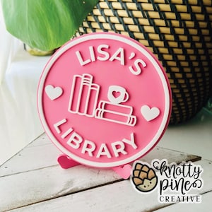 Könnte beinhalten: Ein pinkfarbenes, rundes Schild mit dem weißen Text "LISA'S LIBRARY". Das Schild zeigt weiße Umrisse von Büchern, eine Kaffeetasse mit Herz und Herzen. Es liegt auf einer weißen Holzoberfläche.