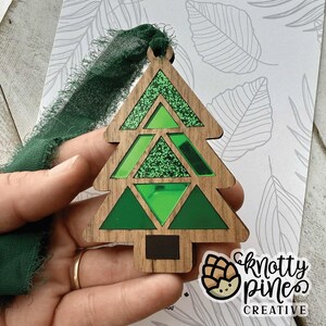 May include: Wooden Christmas tree ornament with green mirrored and glitter accents. The tree has a geometric design and is suspended by a green ribbon. The ornament is held in a hand. The logo for Knotty Pines Creative is in the lower right corner.