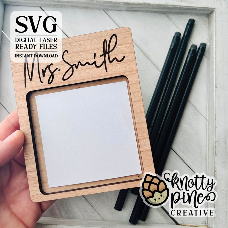 Pu&ograve; includere: Un porta note in legno con un post-it bianco e il testo "Mrs. Smith" inciso sulla parte superiore. Il porta note &egrave; in legno marrone chiaro e ha un piccolo ritaglio per il post-it. Il porta note &egrave; su una superficie bianca con una matita nera.