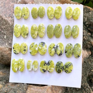 May include: A collection of twenty-eight oval-shaped cabochons arranged in fourteen pairs on a white display card. Each gemstone features a vibrant lime green base color with varying patterns of black and white mineral inclusions throughout the surface.