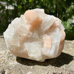 Op de afbeelding: Een cluster van perzik- en witte kristallen. De kristallen hebben een gelaagde uitstraling, waarbij sommige delen doorschijnend lijken. De algemene vorm is afgerond en de kristalformatie is zichtbaar. De steen ligt op een ruw, grijs oppervlak.