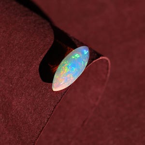 May include: A polished, oval-shaped opal gemstone. The opal displays a milky white base color with iridescent flashes of blue, green, and yellow. The gemstone is set against a deep burgundy background, highlighting its vibrant colors.