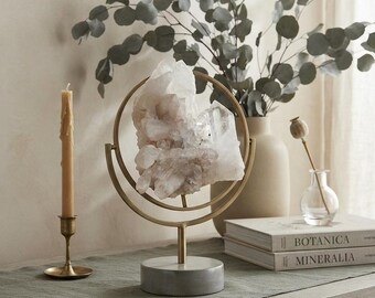 May include: A large, clear, jagged quartz crystal cluster mounted on a gold-toned metal circular stand with a cylindrical gray stone base. The crystal is positioned vertically within the center of the metal ring, creating a decorative display piece.