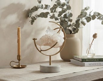 May include: A clear quartz crystal cluster is suspended within a gold-toned metal orbital frame, which is mounted on a circular gray stone base. The display piece is positioned next to a tall, thin, light-colored candle in a brass holder.