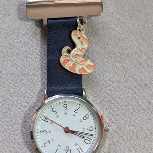 Puede incluir: Un reloj de bolsillo de enfermera plateado con esfera blanca, números negros y segundero rojo. El reloj tiene una correa azul marino y un dije decorativo de serpiente con esmalte naranja y crema. El clip es plateado.