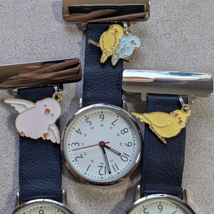 Puede incluir: Tres relojes de solapa plateados con correas texturizadas de color azul oscuro. Cada reloj tiene una esfera analógica blanca con números negros y un segundero rojo. Incluyen dijes de pájaros de esmalte en amarillo, azul y blanco.
