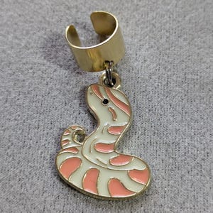 May include: Gold-coloured ear cuff with a dangling snake charm. The snake charm is white with pink and gold accents. The ear cuff is a simple band that wraps around the ear.