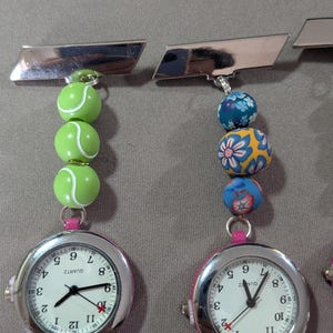 May include: Two silver-tone nurse fob watches with quartz movements and white faces. One watch features three green tennis ball beads, while the other has three blue and yellow floral patterned beads. Both have a rectangular silver pin attachment.