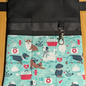 May include: Black canvas utility belt with multiple pockets and a clip for keys. The belt is decorated with a teal fabric featuring a repeating pattern of dogs, cats, and medical symbols.