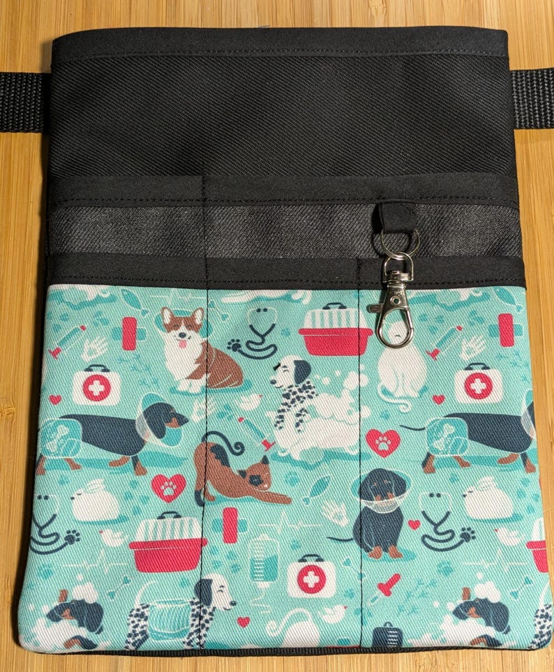 May include: Black canvas utility belt with multiple pockets and a clip for keys. The belt is decorated with a teal fabric featuring a repeating pattern of dogs, cats, and medical symbols.