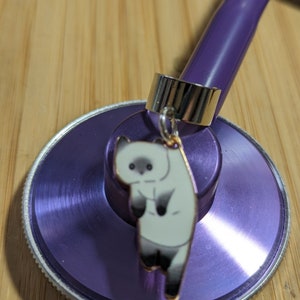 May include: Purple stethoscope with a silver charm attached. The charm is a cartoon white cat with black eyes.