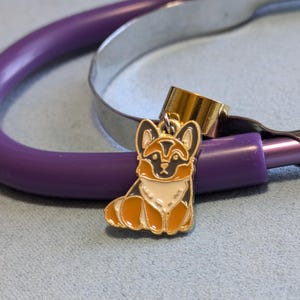 Può includere: Un ciondolo per stetoscopio dorato con un cane pastore tedesco di cartone animato con pelo nero, marrone e bianco. Il ciondolo è su uno stetoscopio viola.