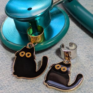 Puede incluir: Un estetoscopio turquesa con un anillo dorado y dos colgantes de gato negro con detalles dorados. Los colgantes de gato tienen ojos amarillos y cuelgan de anillos plateados.