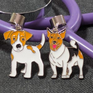Può includere: Due ciondoli in smalto metallico a forma di cane, ciascuno dotato di una contromaglia argentata per l'aggancio. Il ciondolo a sinistra raffigura un cane bianco e arancione, quello a destra un cane bianco, arancione e nero con lingua rosa.