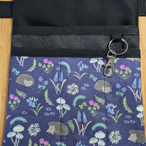 Könnte beinhalten: Eine blaue Stoffschürze mit einem Schlüsselring. Die Schürze hat eine Tasche mit einem blauen Blumenmuster mit Igeln, Pilzen und Blumen. Der obere Teil ist schwarz und die Schürze hat einen schwarzen Riemen.