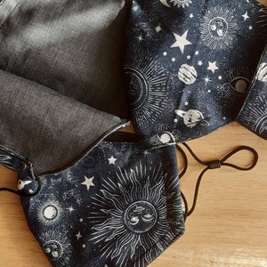 May include: Three black fabric face masks with a white celestial print. The print features suns, moons, stars, and planets. The masks have black elastic ear loops.