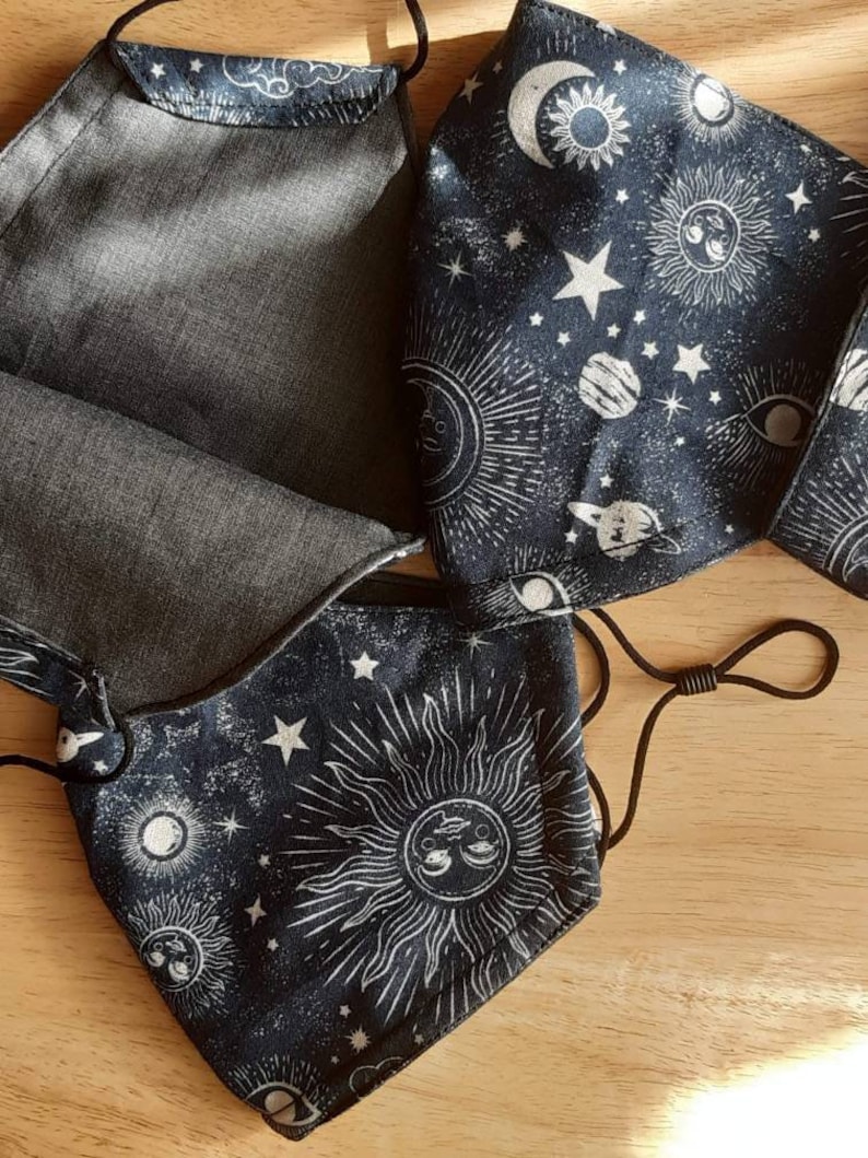 May include: Three black fabric face masks with a white celestial print. The print features suns, moons, stars, and planets. The masks have black elastic ear loops.