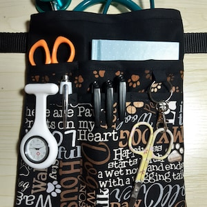 May include: Black fabric utility belt with multiple pockets, designed for veterinary professionals. It features a stethoscope, scissors, a watch, and other medical tools. The fabric has a paw print pattern and text that reads "Dog House, Walk, Paw Prints, Unconditional".