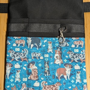 May include: A black canvas tool belt with multiple pockets and a blue fabric lining featuring a pattern of Australian Shepherd dogs wearing cone collars and other medical supplies. The belt has a silver metal clip.