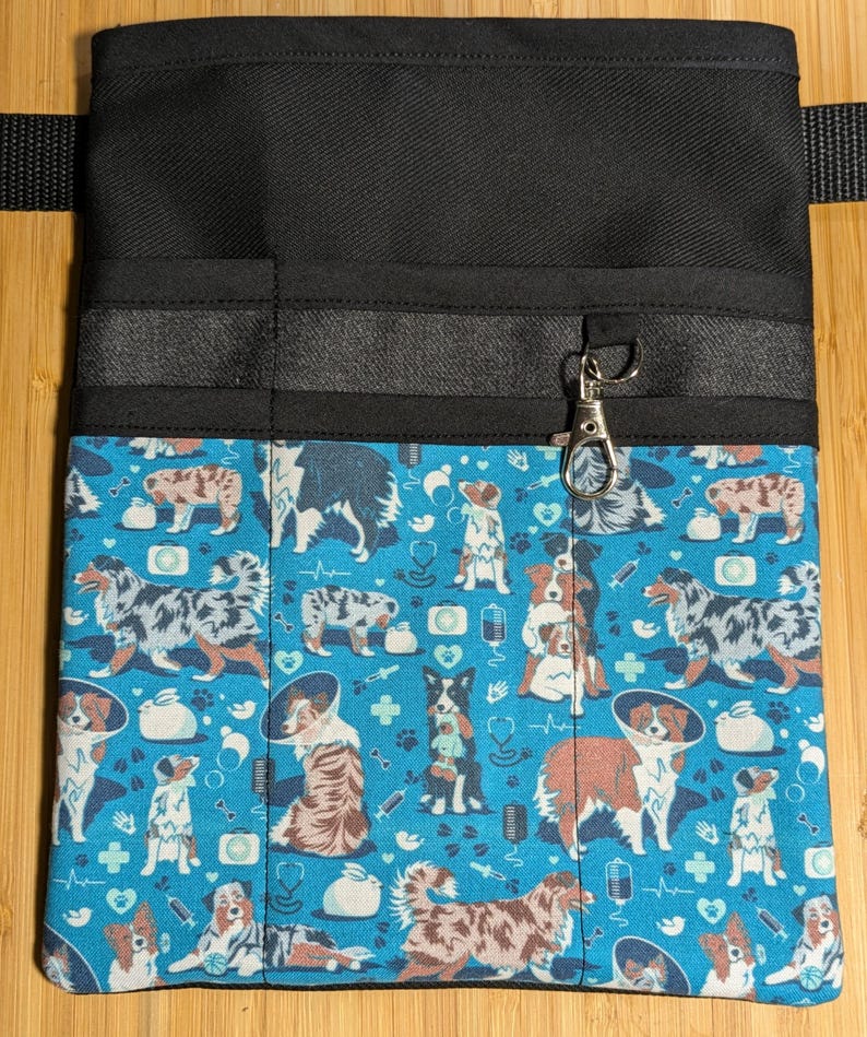 May include: A black canvas tool belt with multiple pockets and a blue fabric lining featuring a pattern of Australian Shepherd dogs wearing cone collars and other medical supplies. The belt has a silver metal clip.