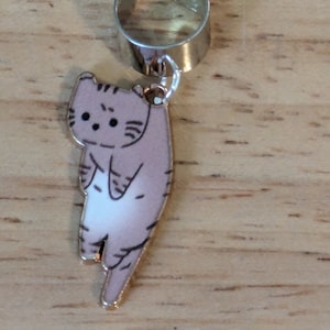 May include: A silver ear cuff with a dangling charm of a cartoon cat with brown and white fur. The cat is facing forward with its back to the viewer.