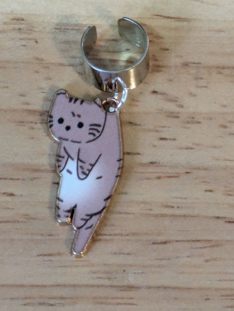 May include: A silver ear cuff with a dangling charm of a cartoon cat with brown and white fur. The cat is facing forward with its back to the viewer.