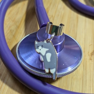 May include: Purple stethoscope with a silver charm of a cat hanging from it. The charm is attached to a silver ring.