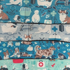 May include: A blue and white fabric with a repeating pattern of dogs, cats, and veterinary-themed symbols. The fabric features a variety of breeds, including a corgi, a dalmatian, a terrier, and a cat. The symbols include a stethoscope, a first aid kit, a bone, and a paw print.