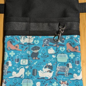 May include: Black canvas utility belt with multiple pockets and a blue and white patterned fabric flap. The flap features a repeating pattern of cats, dogs, bones, and medical symbols. A silver clip is attached to the belt.