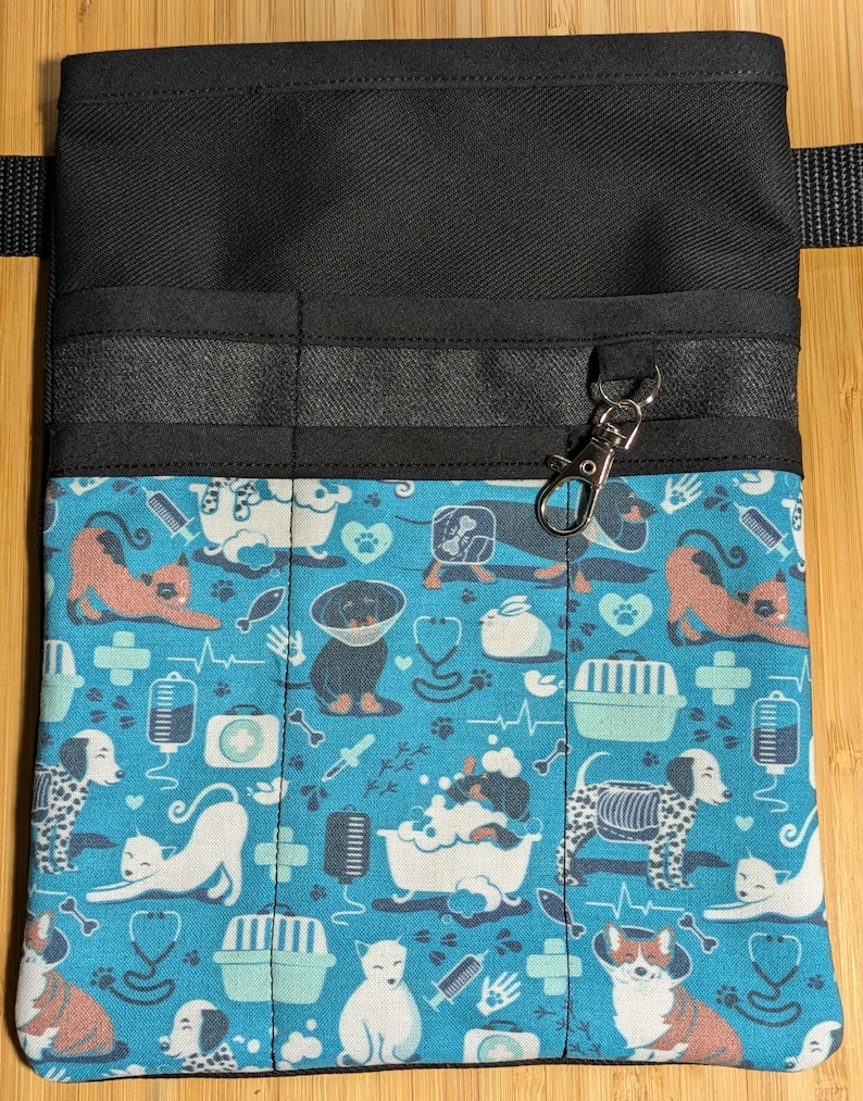 May include: Black canvas utility belt with multiple pockets and a blue and white patterned fabric flap. The flap features a repeating pattern of cats, dogs, bones, and medical symbols. A silver clip is attached to the belt.
