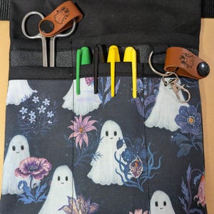 May include: A black utility apron featuring a repeating pattern of white ghosts and colorful flowers. The apron has multiple front pockets containing metal scissors, a green pen, a black pen, two yellow pens, and a brown leather key fob.