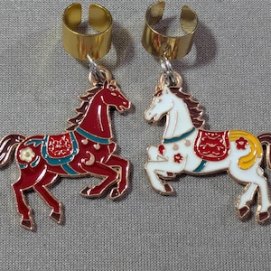 Puede incluir: Dos pendientes tipo ear cuff de metal dorado, cada uno con un dije de caballo colgante. Un caballo es de color rojo oscuro con detalles en verde azulado y blanco, mientras que el otro es blanco con detalles en rojo y amarillo.
