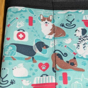 May include: A turquoise blue fabric with a repeating pattern of various dog breeds, including a corgi, dachshund, dalmatian, and a cat. The fabric also features a variety of medical symbols, including a stethoscope, a first aid kit, and a syringe.