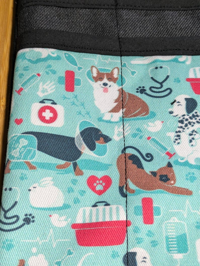 May include: A turquoise blue fabric with a repeating pattern of various dog breeds, including a corgi, dachshund, dalmatian, and a cat. The fabric also features a variety of medical symbols, including a stethoscope, a first aid kit, and a syringe.