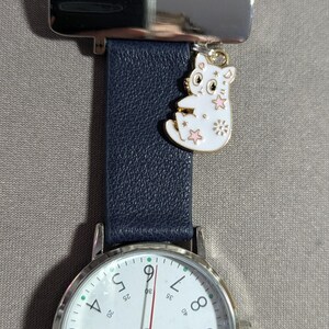 Peut inclure: Une montre argentée avec un bracelet en cuir bleu marine et un cadran blanc. La montre a un charme de chat avec des accents roses et dorés.