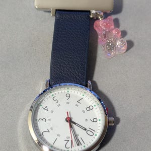 Peut inclure: Une montre de poche d'infirmière argentée avec un cadran blanc, des chiffres noirs et une trotteuse rouge. Elle a un bracelet bleu marine et un charm en forme d'ours en gélatine rose. Le diamètre de la montre est d'environ 3,8 cm.