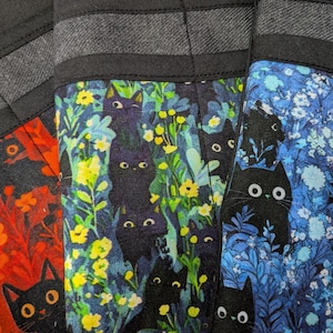 May include: Three fabric panels featuring black cat illustrations amidst colorful floral patterns. The left panel displays orange and red flowers, the center panel shows green and yellow foliage, and the right panel features blue and white blossoms.