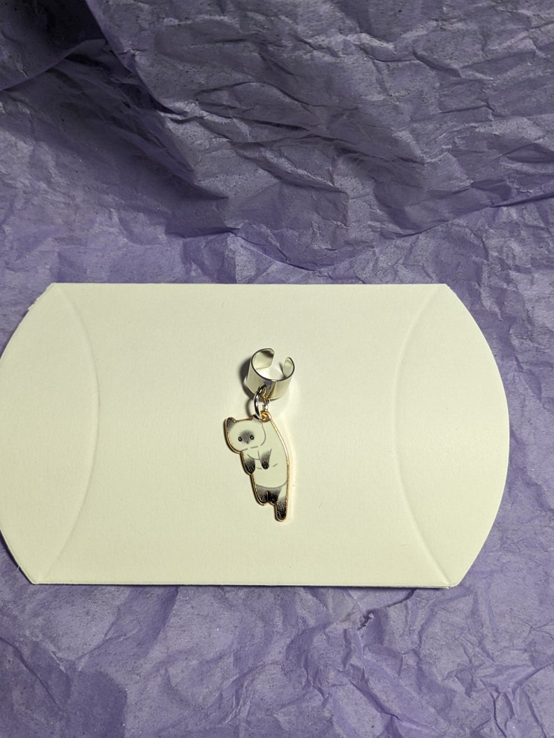 May include: A silver ear cuff with a dangling charm of a white cat with gray markings. The cat is facing to the left and has a surprised expression.
