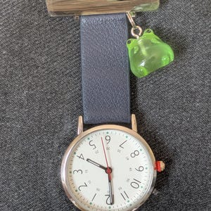 May include: A silver-tone nurse watch with a dark blue strap and a white analogue face featuring black numerals from 1 to 12 and smaller increments of 5. A translucent green frog charm hangs from the top silver pin, and the watch has a red crown.