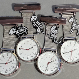 May include: Five silver nurse fob watches with white faces, black numerals, and red second hands. Each watch features a different black and white animal skeleton charm: tapir, parrot, lizard, and horse. The watches have a mesh band and a clip.