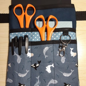 Könnte beinhalten: Ein blauer Stoff-Werkzeuggürtel mit mehreren Taschen, bedruckt mit Narwal- und Orca-Motiven. Der Gürtel hat zwei Paar orangefarbene Scheren, drei schwarze Stifte und einen silbernen Schlüsselanhänger.