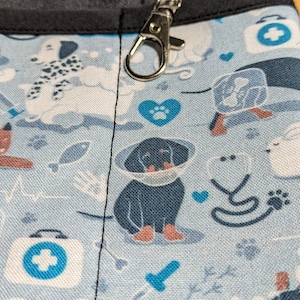 May include: A blue and white fabric with a pattern of dogs, hearts, and medical symbols. A silver metal clip is attached to the top of the fabric.