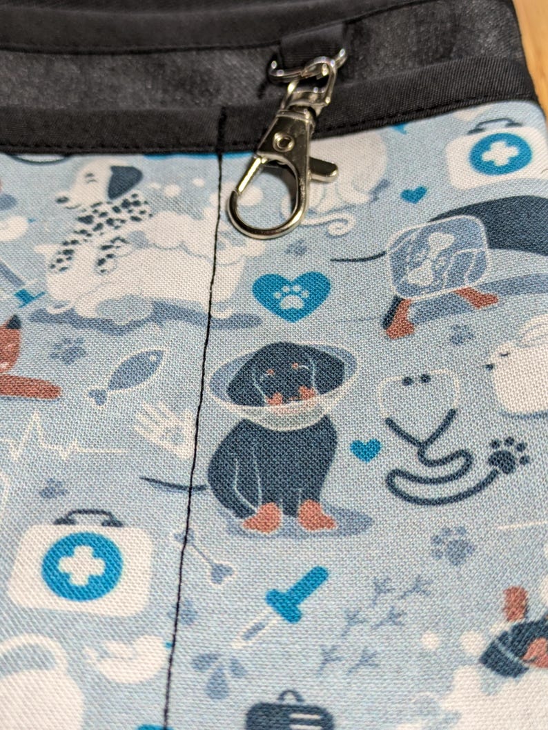 May include: A blue and white fabric with a pattern of dogs, hearts, and medical symbols. A silver metal clip is attached to the top of the fabric.