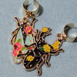 May include: A pair of gold-tone ear cuffs with enamel insect and floral designs. One cuff features a black stag beetle with yellow flowers and green leaves. The other cuff features a blue and green dragonfly with pink and red flowers.