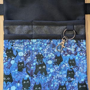 May include: A black fabric utility belt with multiple pockets and a black cat print. The fabric has a blue and white floral pattern. A silver key chain is attached to the belt.