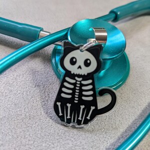 Può includere: Un ciondolo a forma di scheletro di gatto bianco e nero con un anello color argento è attaccato a uno stetoscopio turchese. Il ciondolo ha un design a forma di teschio e gabbia toracica. Lo stetoscopio è turchese e poggia su una superficie grigia.
