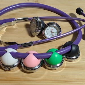 May include: A purple stethoscope with a silver watch and four colored watch fobs: white, pink, green, and black. The watch has a white face with red and black hands. The stethoscope is on a wooden surface.